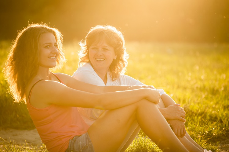 Teenage girl talking with her mother sitting on grass in nature, flare from setting sun in photo