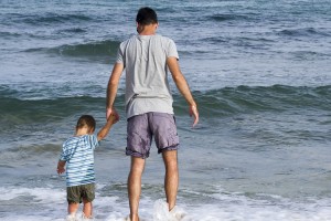 Father and child son stnading on a beach in shallow sea water.