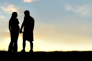 A silhouette of a happy young couple in a relationship, holding hands and talking as they walk outside at sunset.