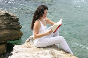 Lady sitting on edge of rock overlooking the ocean and reading a book